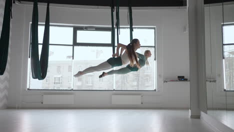 Two-young-yogi-women-doing-aerial-yoga-practice-in-green-hammocks-in-fitness-club.-Beautiful-females-working-out-in-class-performing-aero-yoga.-Variation-of-Parsvottanasana-Pyramid-pose